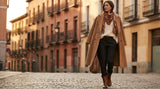 A stylishly layered woman in a camel coat, chunky knit, and silk scarf walks along a cobblestone European street in soft winter light.